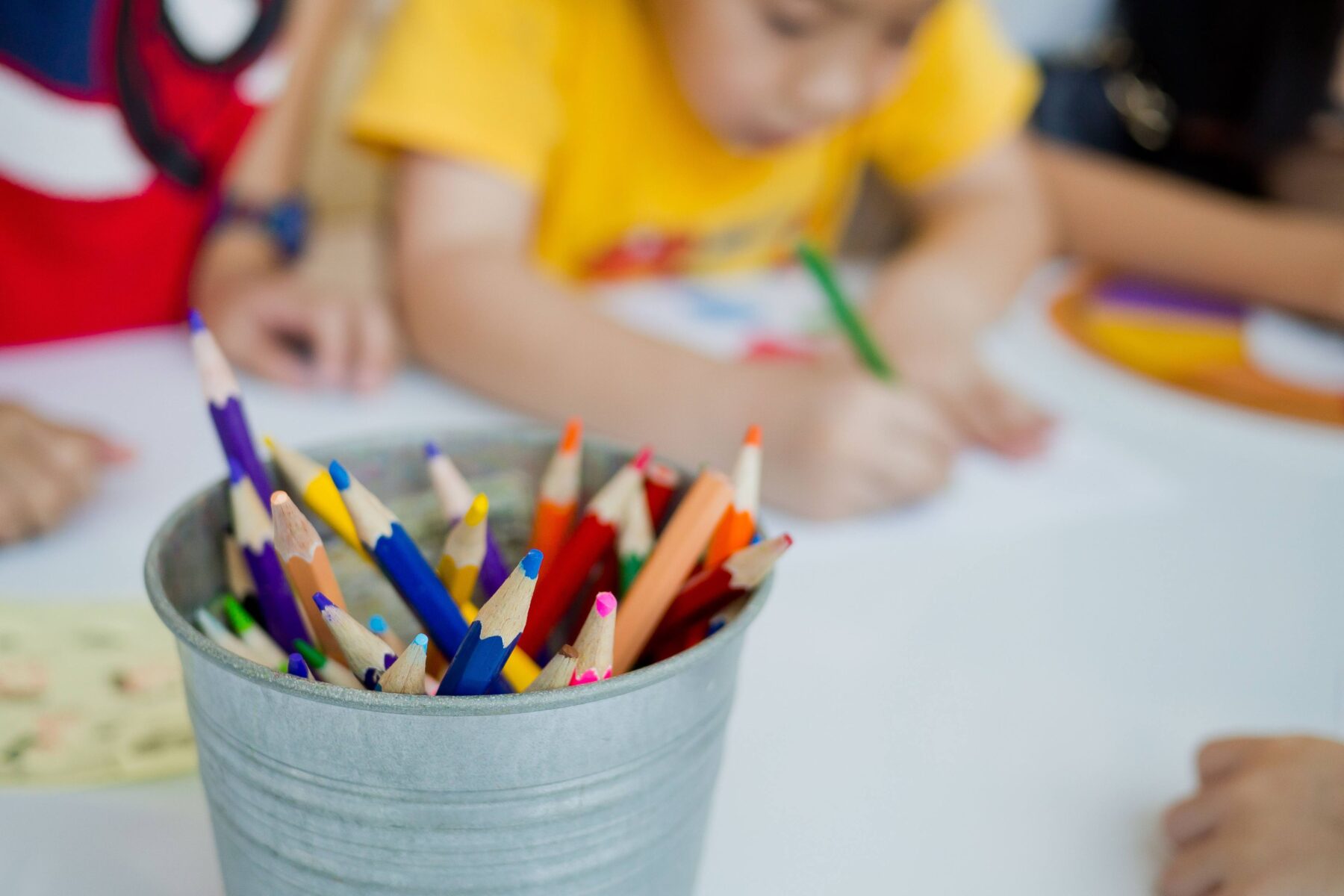 A bucket of colored pencils sitting on a desk, children drawing in the background